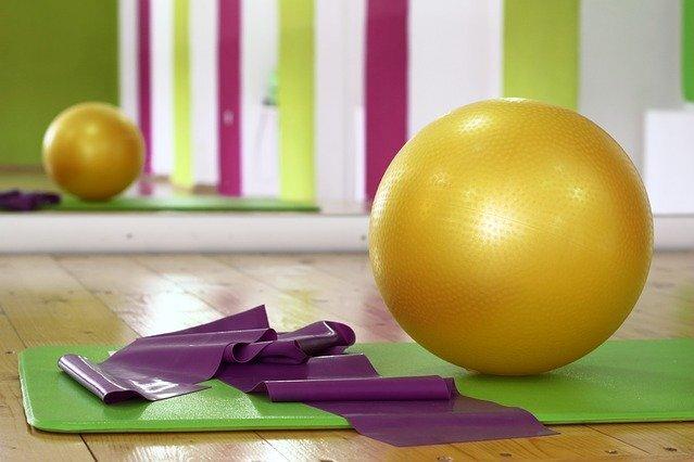 Goldener Gymnastikball und lila Widerstandsbänder auf einer grünen Trainingsmatte in einem Fitnessraum.